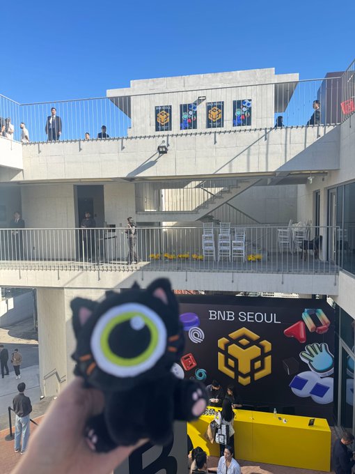 A photo taken at BNB SEOUL event showing a person holding a black cat-shaped plush toy with a green eye in the foreground, with the event venue and BNB branding visible in the background.