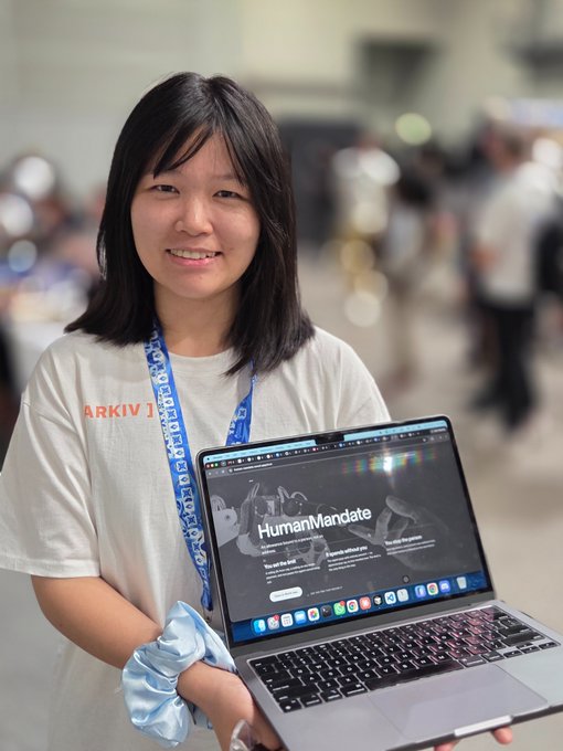 A smiling young woman wearing a lanyard and a t-shirt reading 'ARKIVI' holds up a laptop displaying the 'HumanMandate' website.