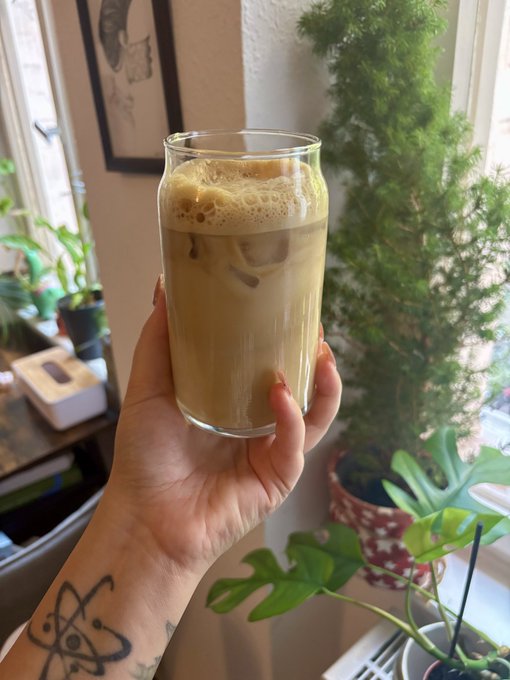 A hand with red nail polish and an eye tattoo on the wrist holds a glass of iced coffee in a bright room filled with potted plants.