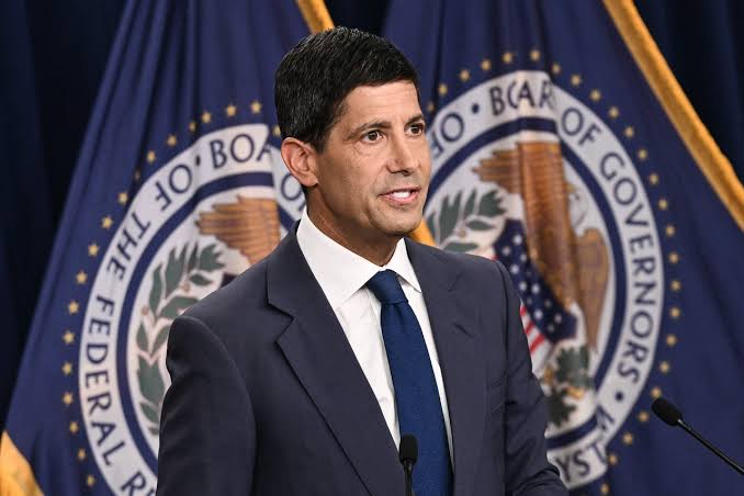 A man in a dark navy suit and blue tie stands at a podium, speaking in front of Federal Reserve Board of Governors seals, illustrating the Fed's hawkish July meeting minutes where rates were held at 3.50%–3.75% despite three dissenters favoring a 25 bps hike amid inflation worries.