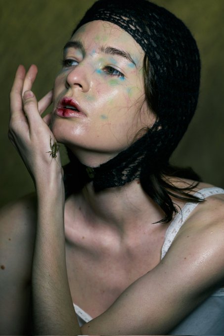 Close-up portrait of a woman with dark hair wearing a black lace headpiece, with colorful paint or makeup splattered on her face, her hand raised near her cheek.