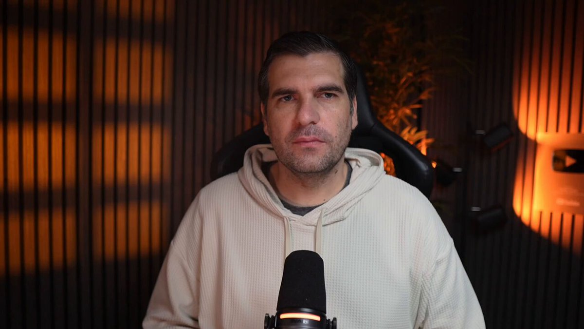 The image shows a man wearing a white hoodie sitting behind a microphone with his hands on his knees. The background features a dark wall with orange lights shining through, creating an interesting contrast between light and shadow. There are also some plants visible in the scene. The photo is related to a post that warns about the challenges faced by Fortune 500 companies due to outdated infrastructure from the 1990s era.