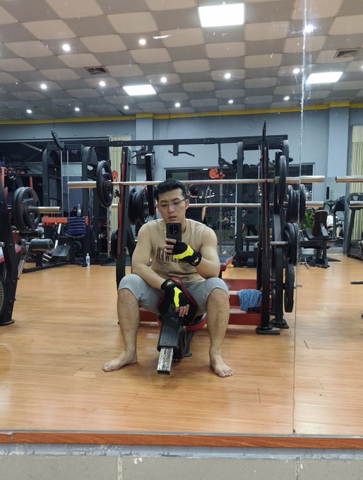 A barefoot man in a sleeveless shirt and weightlifting gloves takes a mirror selfie in a gym, sitting on a bench surrounded by power racks and barbells. The image complements his message about neglecting physical health for years while focused on crypto trading and airdrops, now struggling to lift even half of his former capacity.
