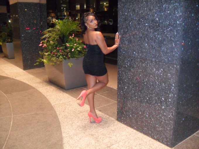 A woman with braided hair wearing a short black dress and pink high heels poses playfully against a dark marble pillar in an upscale indoor setting with planters.
