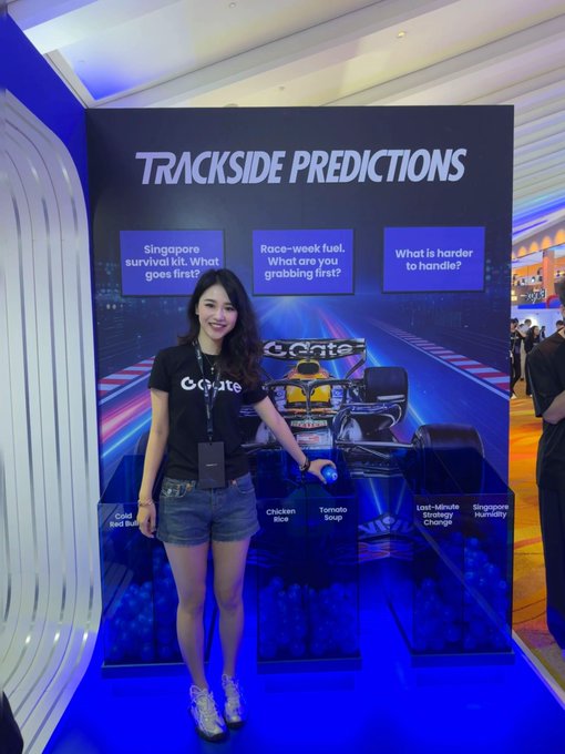 A woman in a GoMate t-shirt and denim shorts poses next to a 'TRACKSIDE PREDICTIONS' display featuring Formula 1 racing questions and answers including 'Chicken Rice' and 'Tomato Soup'.