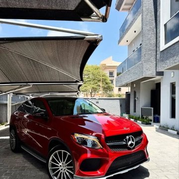 A red Mercedes-Benz SUV is parked on a paved driveway under a dark shade sail in front of a modern multi-story residential building.