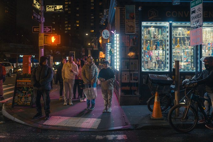 A nighttime street scene showing pedestrians waiting at a crosswalk next to a brightly lit convenience store displaying signs for 'SHARED LANE' and 'SMOKE SHOP'.
