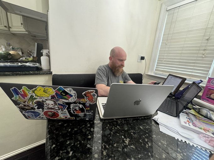 A man with a beard works at a granite kitchen counter cluttered with three laptops and stacks of papers, one laptop covered in stickers including "$FWA." The "I'M FINE" caption paired with the chaotic multi-screen workspace and "$FWA" sticker humorously suggests he is actually overwhelmed, likely working on or stressing over his $FWA investments rather than being fine.