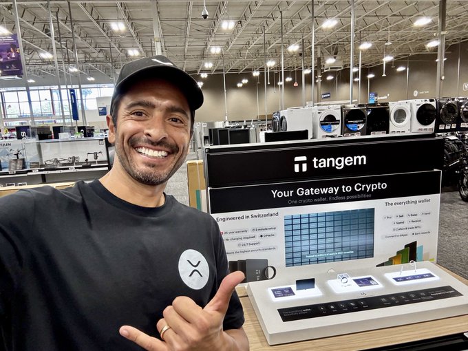 The image features a man wearing a black shirt and hat standing next to a display of electronic devices at a store. The man appears to be posing for a photo, with his hand raised in front of him as if he's giving a thumbs-up gesture or making a peace sign. The background shows various electronic gadgets on shelves, including TVs and laptops. The image also includes text that reads "When the community shows up like this, we show up back to you!".