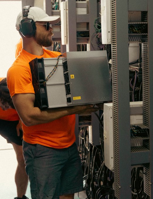 A man wearing a white cap, safety ear protection, sunglasses, and an orange t-shirt carries a server unit while installing it into a server rack filled with tangled cables.