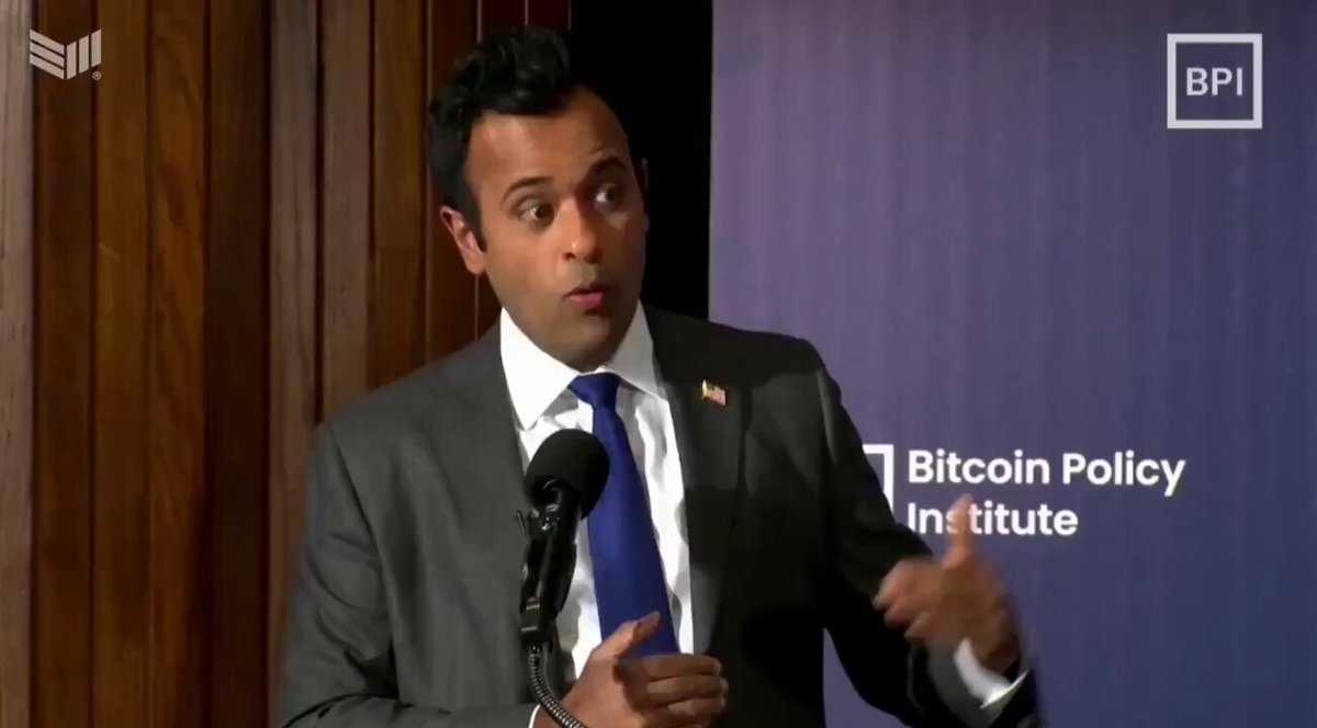 The image shows a man wearing a suit and tie standing at a podium with his hands raised, giving a speech or presentation. The background features a banner that reads "BITCOIN POLICY INSTITUTE" and "VIVEK RAMASWAMY EXPLAINING WHY EVERY COMPANY WILL BUY AND HOLD", suggesting the man is discussing Bitcoin and its potential impact on companies.
