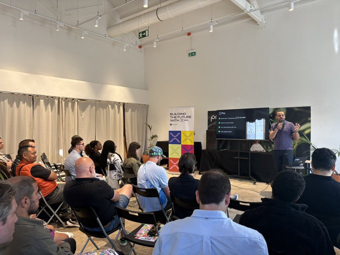 The image shows a man standing at a podium with a whiteboard behind him, giving a presentation or lecture to an audience of people seated in chairs around the room. The attendees appear engaged and attentive as they listen to the speaker's words. The setting appears to be indoors, possibly within a conference room or similar space designed for presentations and discussions.