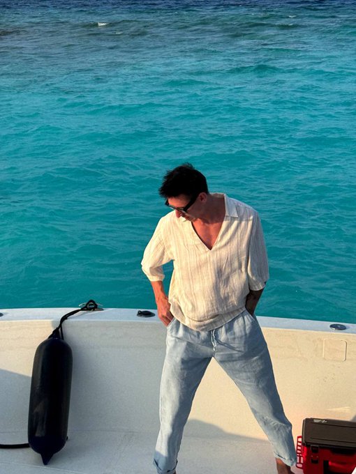A man in sunglasses and a linen shirt stands on a boat in the ocean, looking down. The tranquil, blue water and relaxed attire contrast sharply with the post's theme of cryptocurrency trading losses on Robinhood.