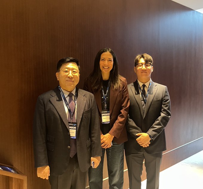 Three professionally dressed individuals pose together against a wooden wall at an event; the two men wear suits and 'VISITOR' lanyards, while the woman in the center wears a brown blazer and a 'SPEAKER' lanyard.