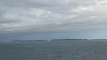 A serene seascape showing two small islands on the horizon under a cloudy, overcast sky over calm ocean water.