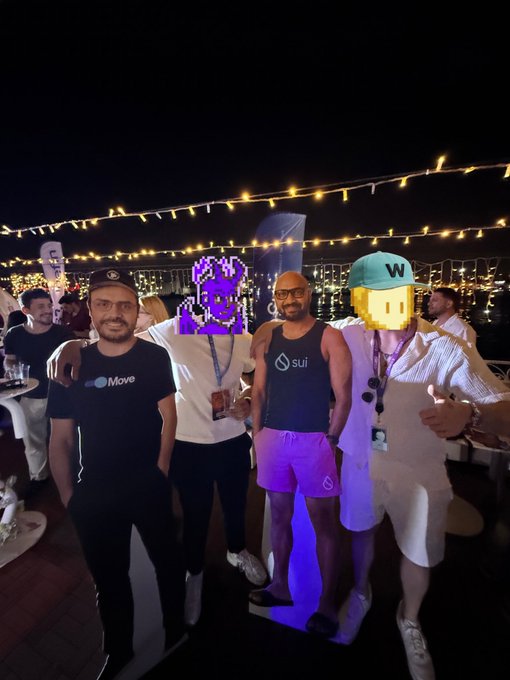 The image shows a group of people posing for a photo at an event or gathering. There are four individuals standing together, with one person wearing a hat that reads "Sui TR community" and another wearing a hat that says "@EmanAbio". The background features string lights, creating a festive atmosphere. The caption to the image includes mentions of the event being held in Turkey and the names of some people involved, such as @SuiTRCommunity and @EmanAbio.