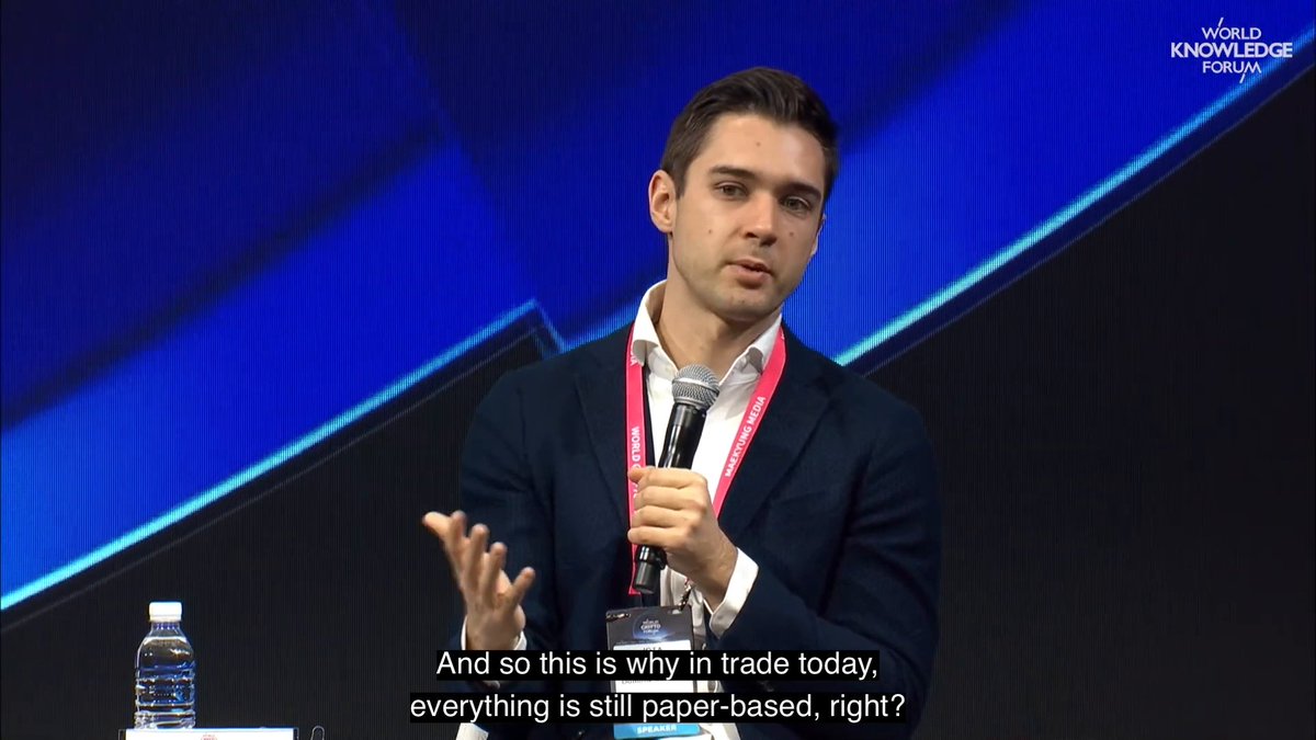 The image shows a man wearing a suit standing behind a microphone, giving a speech or presentation. The background of the image features a blue wall with white text that reads "Focus on what is in the image and how it relates to the post." This suggests that the image might be related to a discussion about the challenges faced by businesses in today's global trade environment.
