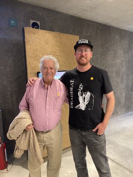 A man in a black "DEADBEAT" t-shirt and a man in a pink button-down shirt stand smiling for a photo indoors.