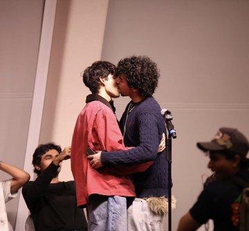 The image shows two men kissing passionately, with one man wearing a red shirt and the other wearing a blue sweater. In the background, there are three more people present, but they are blurred out and not the main focus of the photo. The caption "Monad son or berabaddie daughter" suggests that this scene might be related to a discussion about the concept of monad, which is a form of Islamic thought.

The image also contains some text elements such as "post" and "Berabaddie", but they are not directly related to the main subject matter. The focus remains on the kissing couple in the foreground, with the additional people in the background serving as an interesting backdrop for this intimate moment.