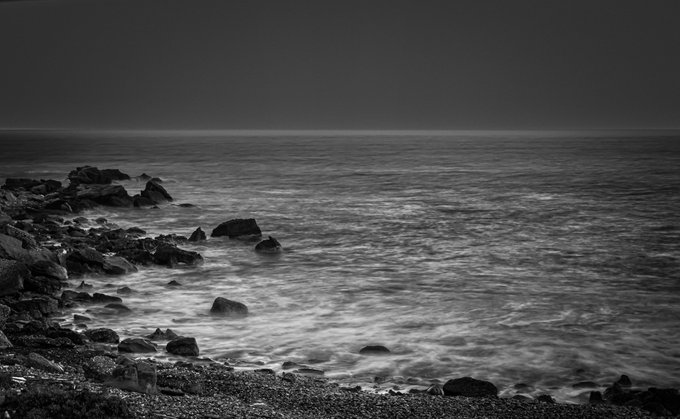 A black and white long-exposure photograph captures a rocky shoreline with misty, motion-blurred waves under a dark sky.