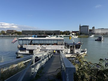 A view from a pier showing two tour boats docked on a river, with a city skyline and historic buildings visible across the water under a clear blue sky.