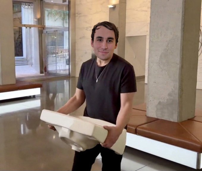 The image shows a man standing in an empty room, holding a large white object or box. He appears to be smiling for the camera while posing with the item he's carrying. The background of the photo features a concrete wall and a bench, which are located near the center of the frame. There is also a text overlay that reads "reminder that this happened 1 year ago and it' s been downhill ever since".