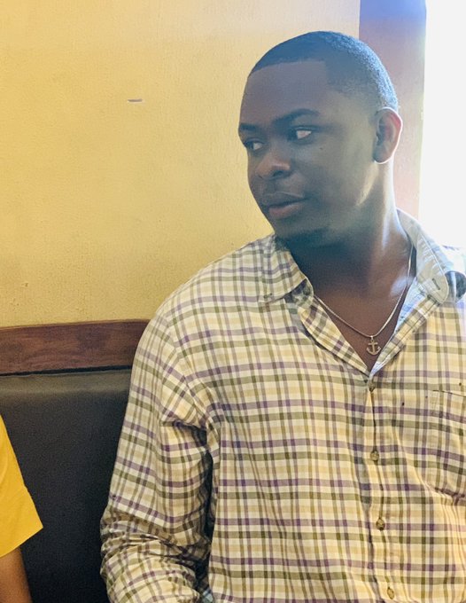 A man in a yellow plaid shirt and an ankh necklace looks to the side while sitting indoors.