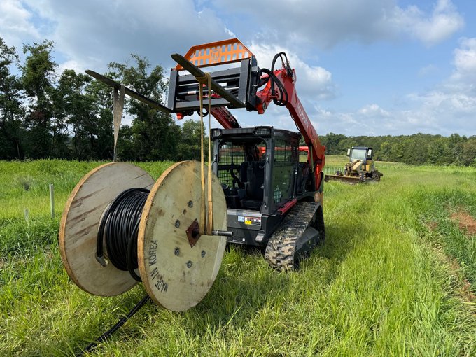 A red tracked skid-steer loader in a grassy field uses a specialized attachment to lift and guide a large wooden cable spool wound with black wire, with a bulldozer visible in the background.