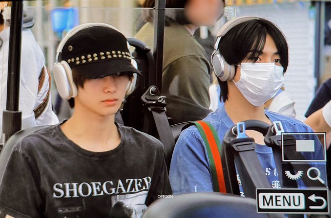 Two young men sit inside a vehicle, likely an airport shuttle; the one on the left wears a studded black cap and a 'SHOEGAZER' t-shirt, while the one on the right wears a blue shirt and a white face mask, both with large headphones.