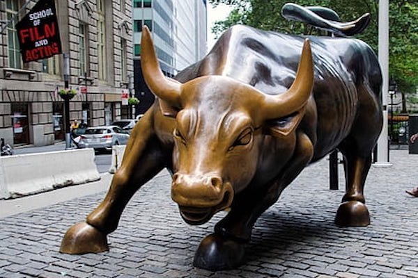 The iconic bronze Charging Bull statue stands on a cobblestone street in New York City, with a 'School of Film and Acting' sign visible in the background.