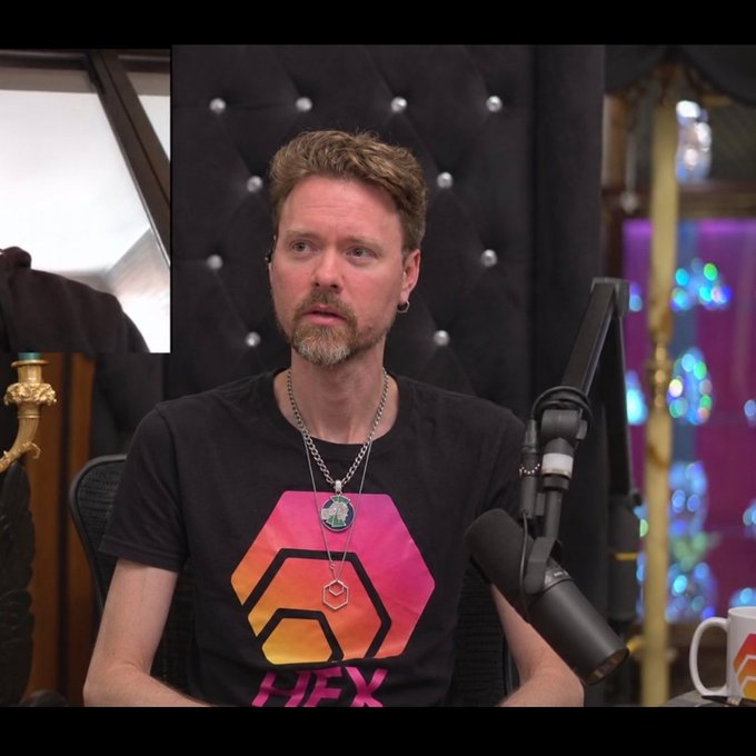 A man with reddish-blonde hair and a goatee sits in a studio setting wearing a black t-shirt with a Hex logo design, positioned in front of a microphone.