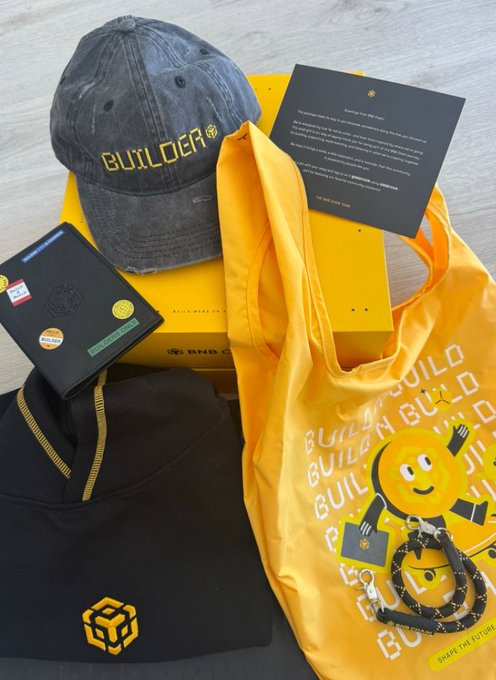 A collection of merchandise branded with 'BUILDER' and 'BNB CHAIN' logos, including a denim cap, a yellow tote bag, a hoodie, a journal, and a keychain with a smiley face, arranged on a surface.