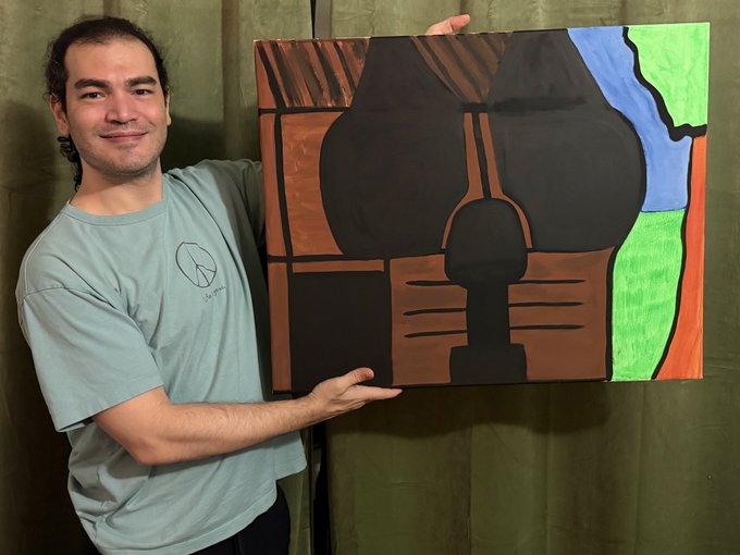 A smiling man in a light blue t-shirt holds up a painting of a stylized skull/animal face in black, brown, green, and blue colors.