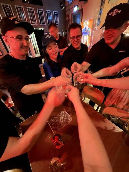 A group of smiling friends toasting with shot glasses around a wooden table, while one person in a black "BORED APE YACHT CLUB" cap (with the recognizable BAYC logo) raises their glass. The scene captures a celebratory moment that ties directly to the post's warm welcome to the Bored Ape Yacht Club community.