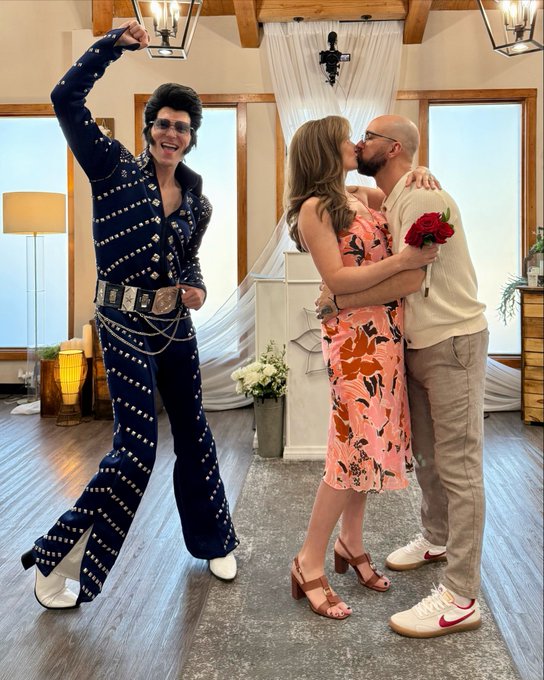 A person dressed as Elvis stands with arm raised next to a couple kissing at a wedding, the woman holding a red rose.