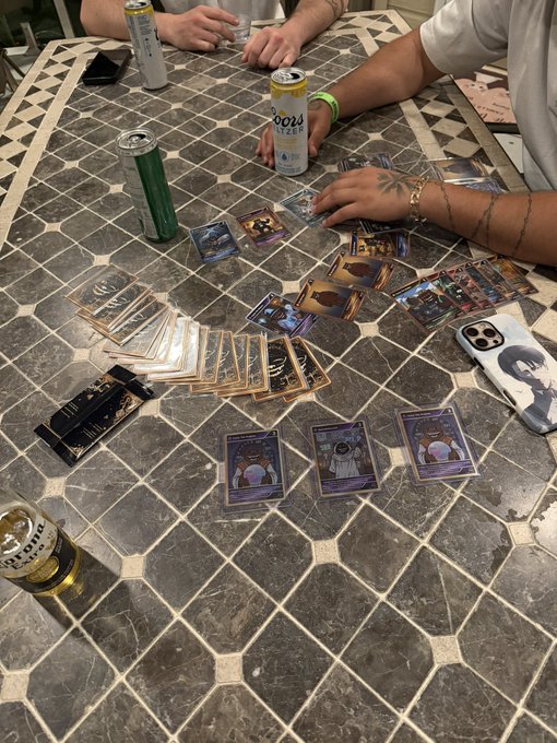 A group of friends gathers around a tiled table with drinks in hand, sorting and laying out FUZZY trading cards as they learn the basics of the collectible card game. Visible cards include "Bullet Bambi" and "Nunzio the Intern," alongside cans of Coors Light and a beer glass, capturing a casual game night atmosphere.