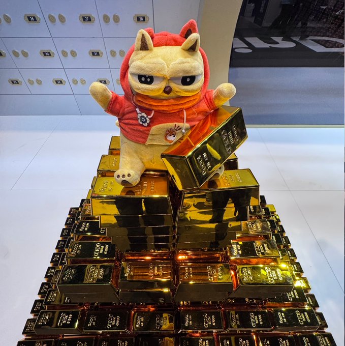 A plush Shiba Inu mascot wearing a red hoodie perched atop a pyramid of stacked gold bars marked "FINE GOLD 999.9," appearing to be a display in a vault room with safety deposit boxes in the background.
