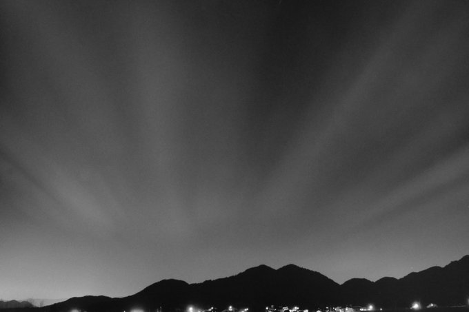 A black and white nighttime photograph captures beams of light radiating upward into a dark sky above a silhouetted mountain range and distant city lights.