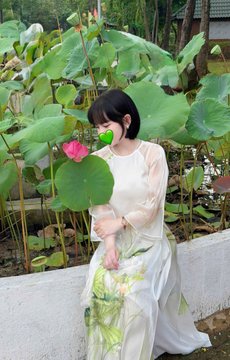 A woman in a flowing cream-colored dress with lotus motifs poses beside a pond filled with vibrant lotus leaves and a pink lotus bloom, evoking the serene elegance of a traditional East Asian summer scene that complements the graceful "Xin chào" greeting to the @0xAlphaGEMs community.