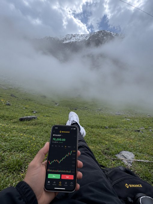 Relaxing on a misty mountainside, someone holds up a phone displaying the Binance app with BTC/USDT at $78,000.00, fittingly trading under the "Binance" branded pants—perhaps a subtle hint about the location being somewhere Binance-affiliated, though the soaring peak and sea of clouds remain a mystery.