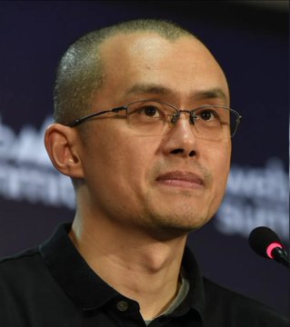 A close-up portrait of a man with short hair and glasses, appearing at what looks like a speaking event with a microphone visible.
