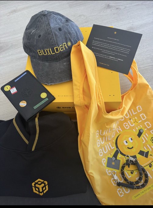 An assortment of merchandise includes a dark cap with 'BUILDER' in yellow text, a yellow tote bag, a black garment with a cube logo, a wallet, and a yellow box.