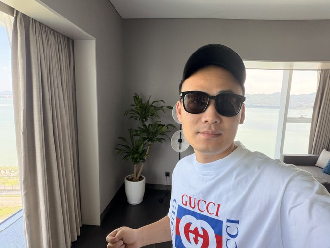 The image features a man wearing sunglasses, a white t-shirt with a logo, and a baseball cap. He appears to be posing for a photo while standing near a window overlooking a body of water. The background includes curtains and a potted plant, adding an element of natural beauty to the scene. The post caption reads "市场行情瞬息万变", which suggests that the man is in a city or urban setting with a focus on architecture and design elements.