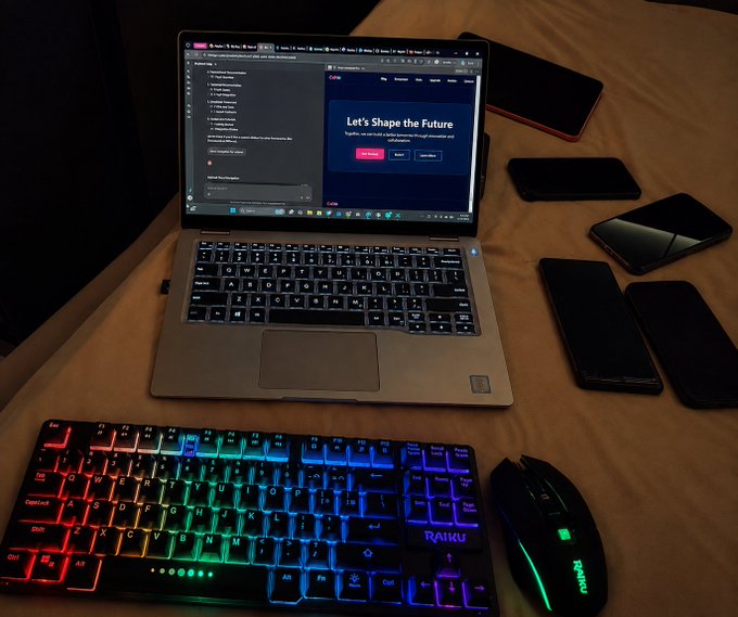 A laptop displaying a ChatGPT interface with the message "Let's Shape the Future" sits on a bed alongside a rainbow-backlit mechanical keyboard (Raiju), a gaming mouse, and several smartphones. The setup humorously illustrates the post's "lazy but have to work" sentiment, showing the classic scene of someone who has moved their entire workstation to bed to stay comfortable while being productive.