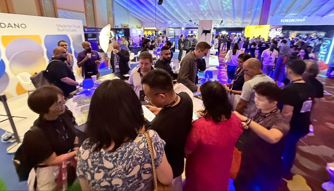 A photograph of a crowded conference or exhibition hall, with numerous attendees gathered around tables and booths, including one labeled 'DANO'.