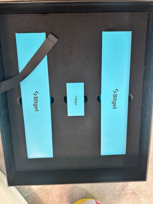 A Bitget-branded gift box containing two teal "Bitget" boxes and a smaller central packet, accompanied by a black strap. This unboxing reveals the contents of the 8th-anniversary merchandise package from Bitget, which the user describes as a surprisingly large and practical gift set.