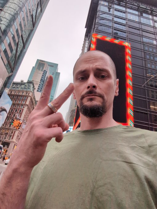 A selfie of a man with a goatee in a green shirt making a peace sign, taken in an urban setting with tall buildings and a large illuminated billboard in the background.