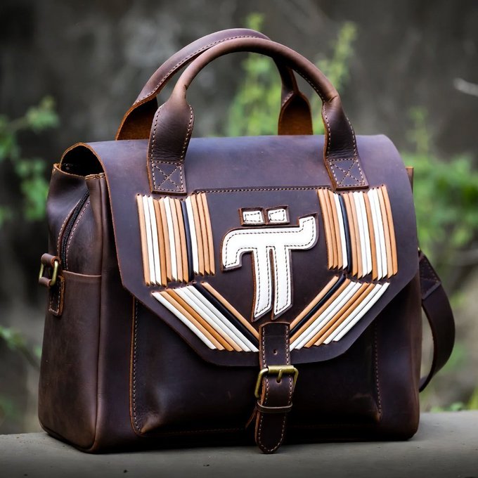 A brown leather messenger bag with a decorative flap featuring a stylized Pi (π) emblem surrounded by a colorful sunburst pattern of layered leather strips.