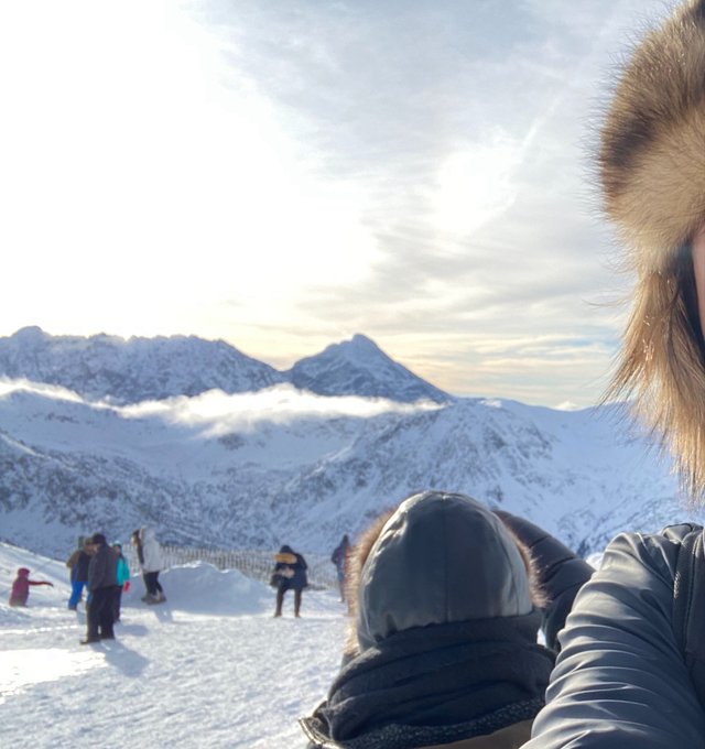 A selfie taken in a snowy mountain landscape shows a person in a furry hat, with other tourists visible on the slope below and peaks rising above clouds in the distance.