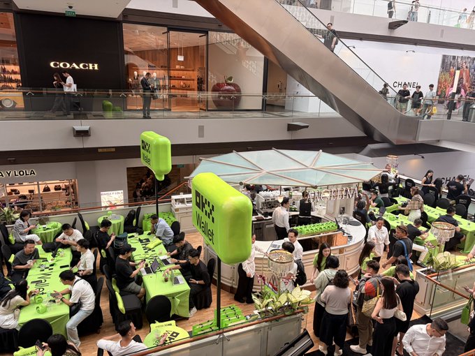 Crowded indoor shopping mall atrium featuring a bright green "Luckin Coffee" promotional pop-up with neon green chairs, surrounded by shoppers and adjacent stores including Coach and Chanel.